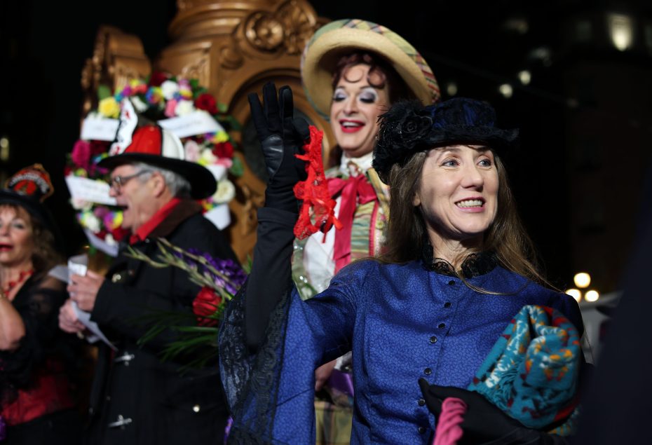 A woman in a blue vintage dress and black hat waves, holding flowers, with people in colorful, elaborate costumes in the background at a nighttime event commemorating the 1906 earthquake.