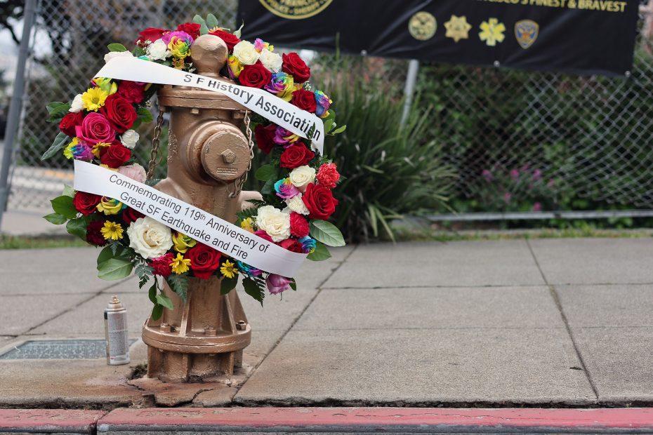 A gold fire hydrant decorated with a colorful floral wreath and banner commemorates the 119th anniversary of the 1906 earthquake and fire. A spray paint can sits nearby on the sidewalk.