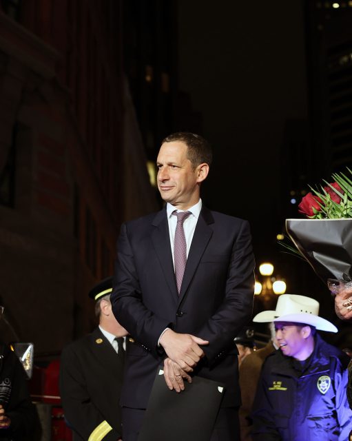 A man in a suit stands outdoors at night, holding a folder amid people in uniforms and a bouquet of flowers, possibly reflecting on the aftermath of the 1906 earthquake.