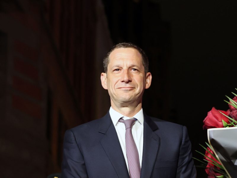 A man in a navy suit and tie stands indoors near a bouquet of red flowers, facing the camera and smiling slightly, as if attending a ceremony commemorating the 1906 earthquake.