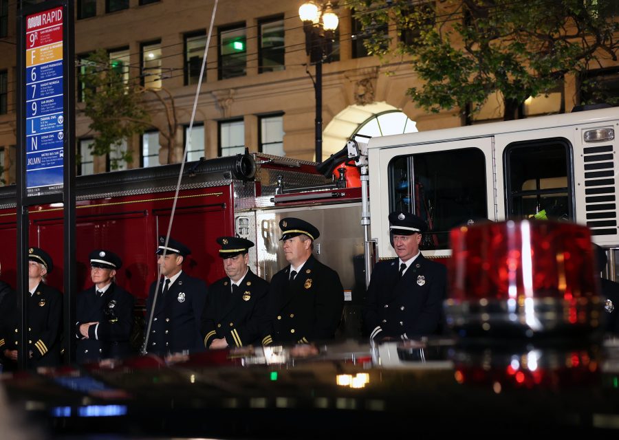 A group of uniformed firefighters stand in front of fire trucks at night, with a public transit sign and building visible in the background, reminiscent of emergency crews responding after the 1906 earthquake.