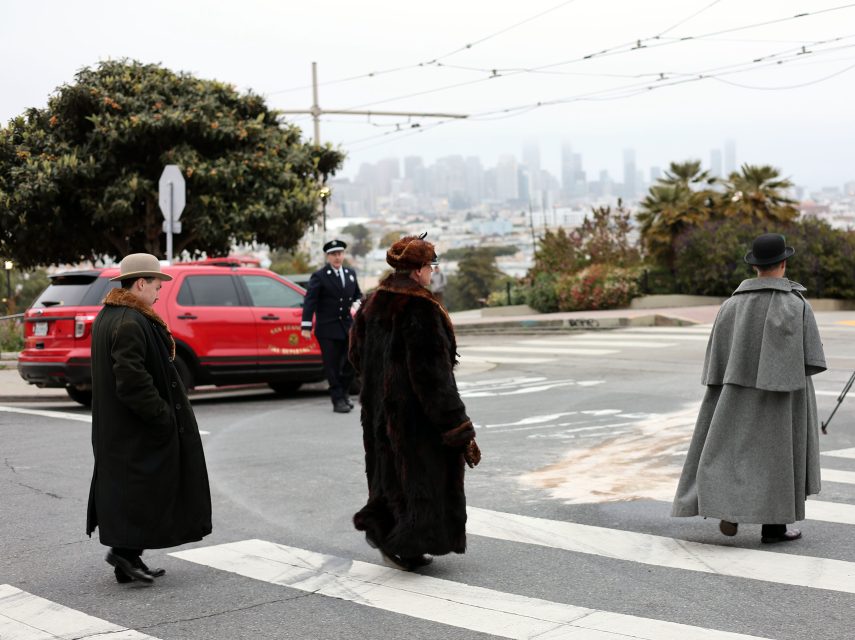 Three people in vintage coats cross a city street at a crosswalk, their style reminiscent of the era surrounding the 1906 earthquake, while a police officer and a red car appear in the background.