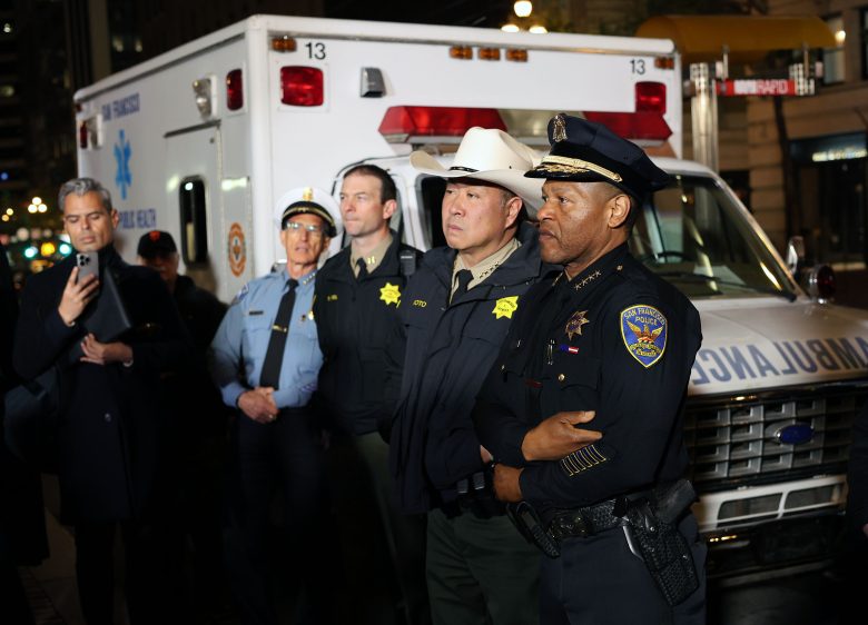 A group of uniformed police officers and officials stand in front of an ambulance at night in an urban setting, reminiscent of emergency responses during the 1906 earthquake.