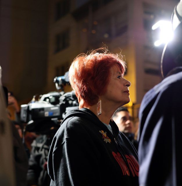 A woman with short red hair stands in profile at night, facing a light source, reminiscent of a scene after the 1906 earthquake, with people and a video camera in the background.
