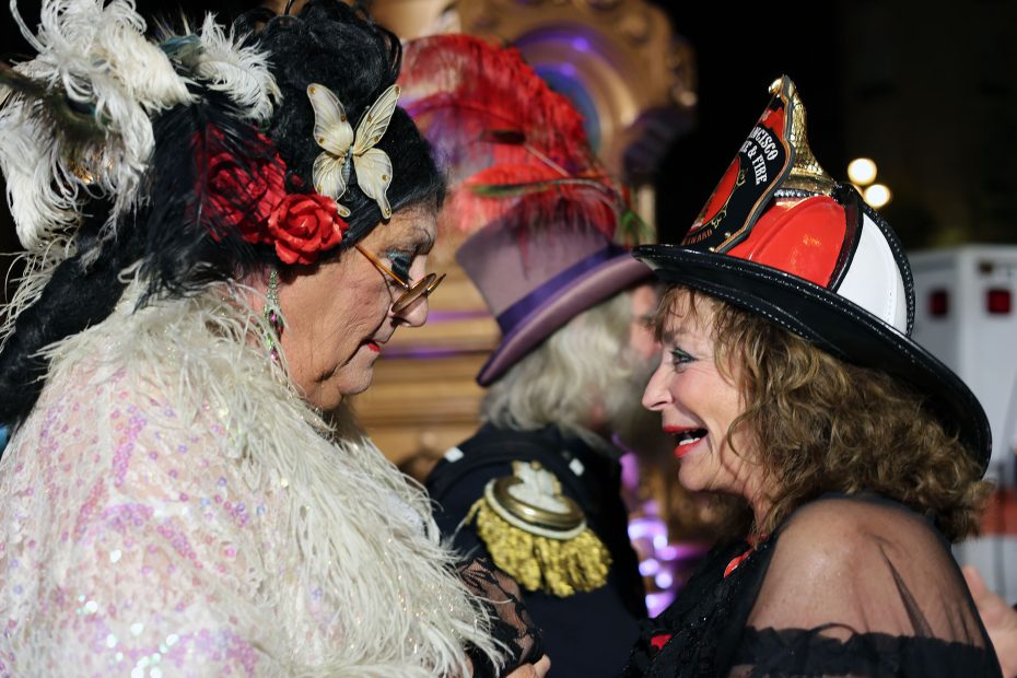 Two people in elaborate costumes and hats stand face to face at an event inspired by the 1906 earthquake, with a person in a decorated jacket in the background.