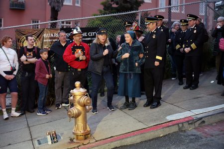 A group of people, including uniformed officials and civilians, stand gathered around a fire hydrant on a city sidewalk near a fence, possibly assessing damage after the 1906 earthquake.