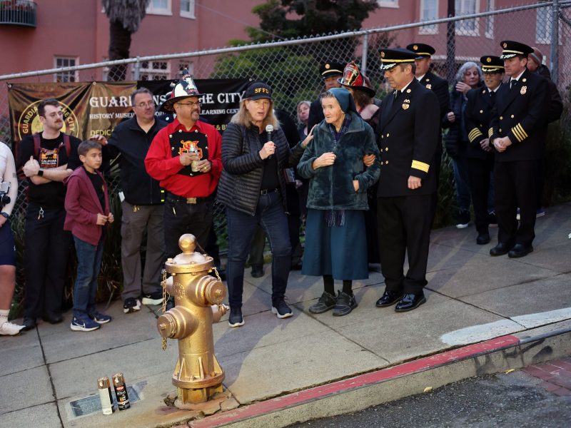 A group of people, including uniformed officials and civilians, stand gathered around a fire hydrant on a city sidewalk near a fence, possibly assessing damage after the 1906 earthquake.