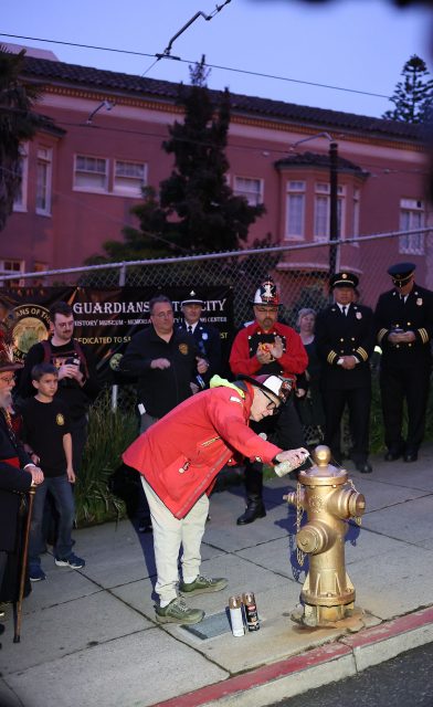 A person in a red jacket and fire helmet paints a gold fire hydrant on the sidewalk, commemorating the 1906 earthquake, while a group of people, including uniformed officials, watch and applaud.