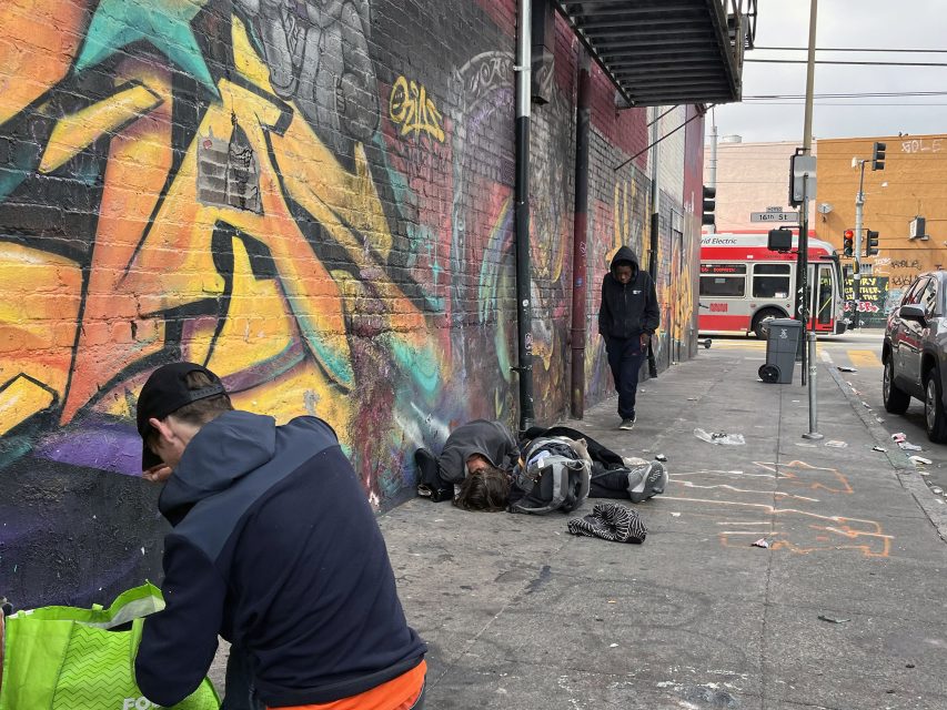 Three people are on a graffiti-covered sidewalk near Kid Power Park; one is bent over a green bag, another lies on the ground, and someone walks by. A city bus and street signs appear in the background.