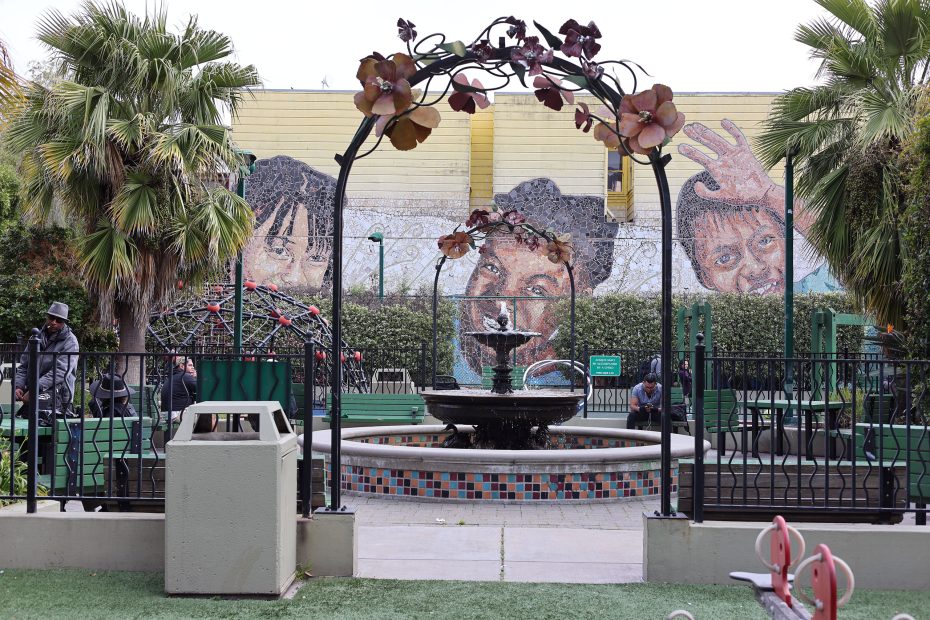 A small urban park features a central fountain, a mosaic mural of faces on a yellow building, palm trees, benches, and people sitting and talking.