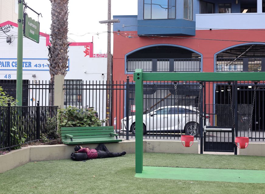 A person lies on the ground near a green bench in a small playground area with swings, adjacent to a fenced street and parked cars.
