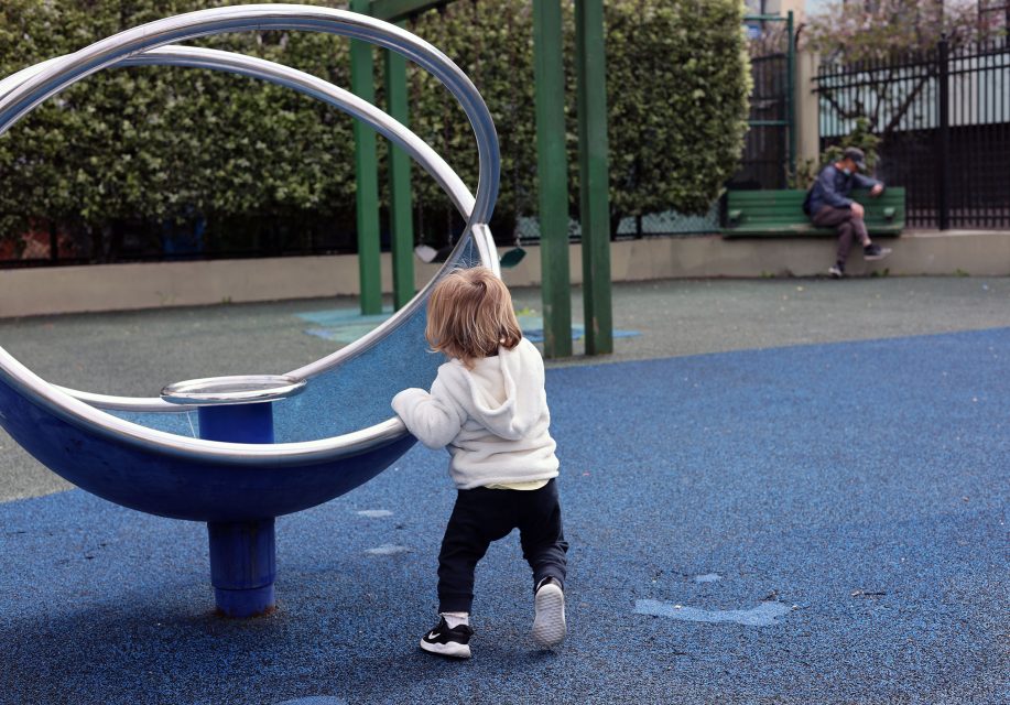 A young child pushes a circular playground structure on blue rubber flooring, while an adult sits on a bench in the background.