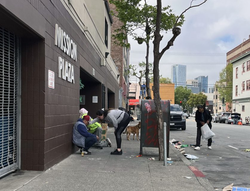 People gather on a sidewalk near Mission Plaza and kid power park as others pick up litter; a dog is present, with tall buildings visible in the background.