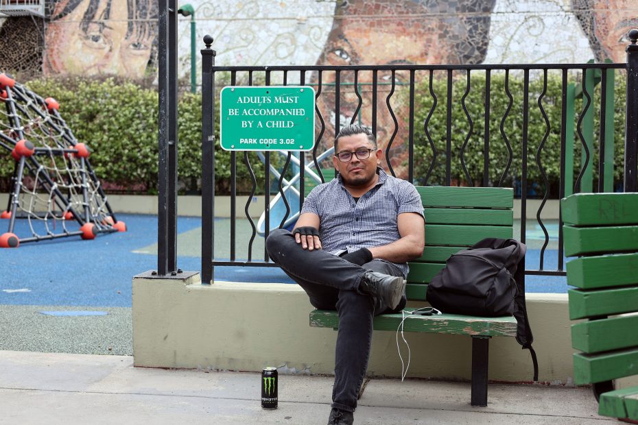 A man sits on a green bench near a playground, with a backpack, headphones, and an energy drink beside him. A sign behind reads “ADULTS MUST BE ACCOMPANIED BY A CHILD.”.
