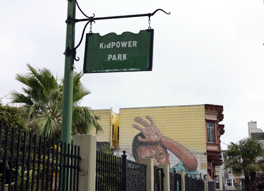 A green sign reading “KidPOWER PARK” hangs near a black fence, with a mural of a child’s face and hand visible on a yellow building in the background.