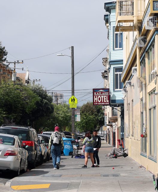 Three people walk on a city sidewalk beside parked cars and buildings, including a hotel with a visible sign; street trees, a pedestrian crossing sign, and nearby Kid Power Park add to the urban scene.