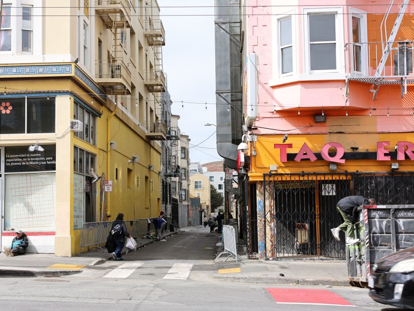 A narrow city alley between yellow and pink buildings, with people collecting items near a garbage bin and debris on the sidewalk, leads toward the lively atmosphere of Kid Power Park nearby.