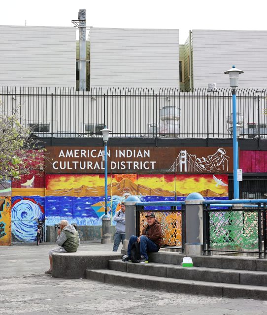 Several people sit near steps in front of a colorful mural labeled “American Indian Cultural District” at Kid Power Park, with a modern building and fence in the background.