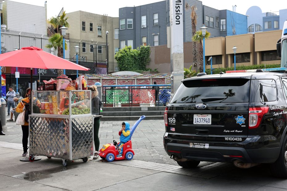 A street vendor stands by a fruit cart under a red umbrella while a child shows kid power in a toy car near a parked police SUV in an urban setting.