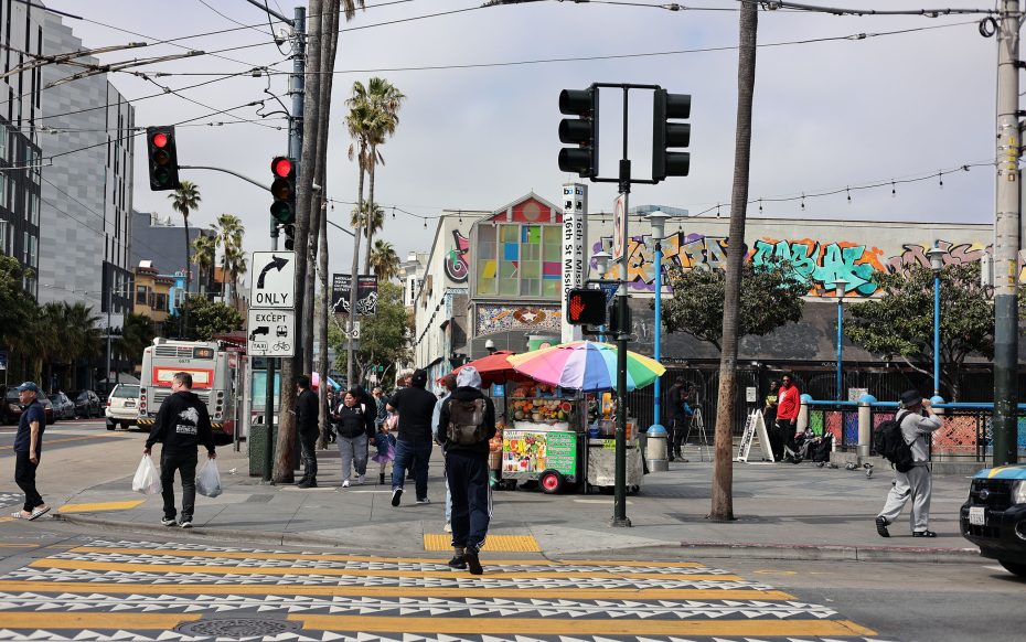 People cross a city street near a colorful food cart, with a graffiti-covered building and palm trees in the background. The traffic light is red, and the lively scene hints at the vibrant spirit of kid power park nearby.
