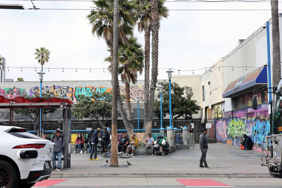 People gather near palm trees and colorful graffiti walls at Kid Power Park in an urban outdoor area; parked and moving cars are visible in the foreground.