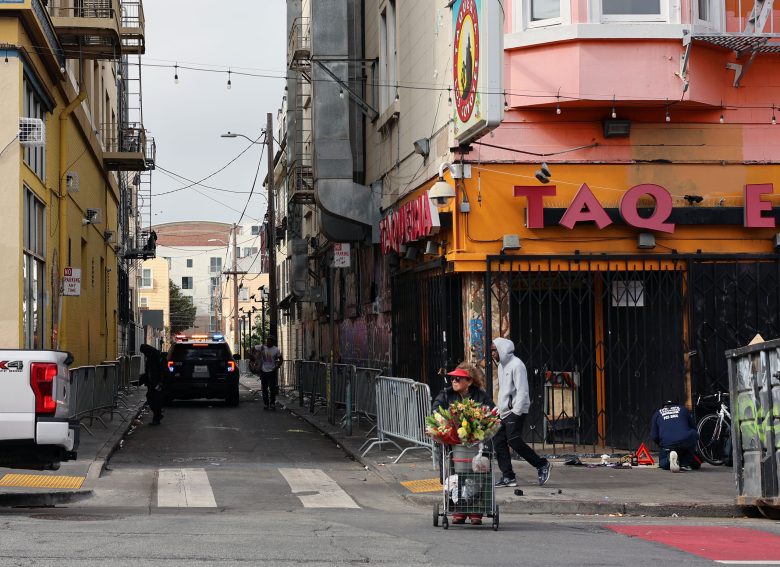 A person pushes a cart filled with flowers past a closed storefront on a city street, while another person in a hooded jacket walks nearby and a police car is parked in the alley.