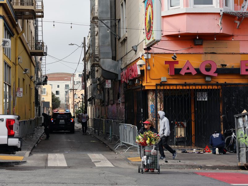 A person pushes a cart filled with flowers past a closed storefront on a city street, while another person in a hooded jacket walks nearby and a police car is parked in the alley.