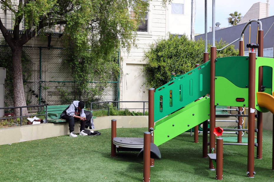 Two people sit together on a bench near a green playground structure in an outdoor park area with grass and trees.
