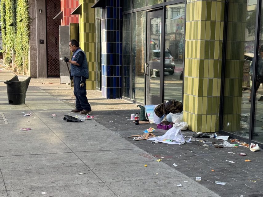 A person is sitting on the ground surrounded by scattered trash near a building entrance on 16th, while another person stands nearby on the sidewalk.