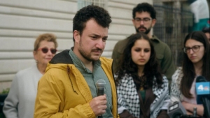 A man in a yellow jacket holds a microphone while standing in front of a group of people outdoors, some looking serious.