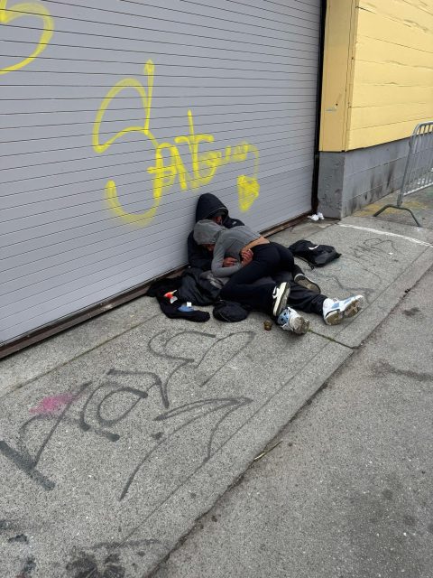 Two people lie on a sidewalk against a garage door with yellow graffiti, surrounded by litter and wearing dark clothing.