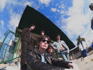 Five people stand and sit outside a building with green railings under a blue sky with clouds; one person in front wears sunglasses and a black leather jacket.