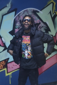 A man wearing sunglasses, a black puffer jacket, and a graphic t-shirt stands in front of a colorful graffiti wall, smiling with his mouth open.