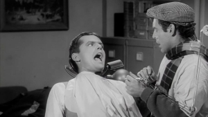 A man sits in a barber chair looking alarmed while another man, dressed casually, holds barber tools and leans toward him.