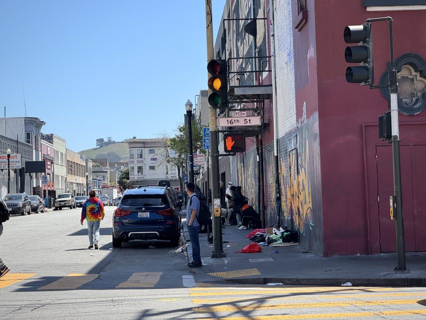 People stand and walk near a busy intersection at 16th Street. Some individuals are gathered by a building with graffiti and scattered belongings on the sidewalk.