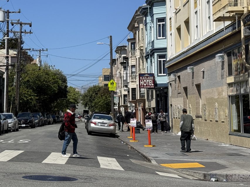 A man crosses a city street at a crosswalk near parked cars and a group of people standing outside a hotel under clear blue skies.