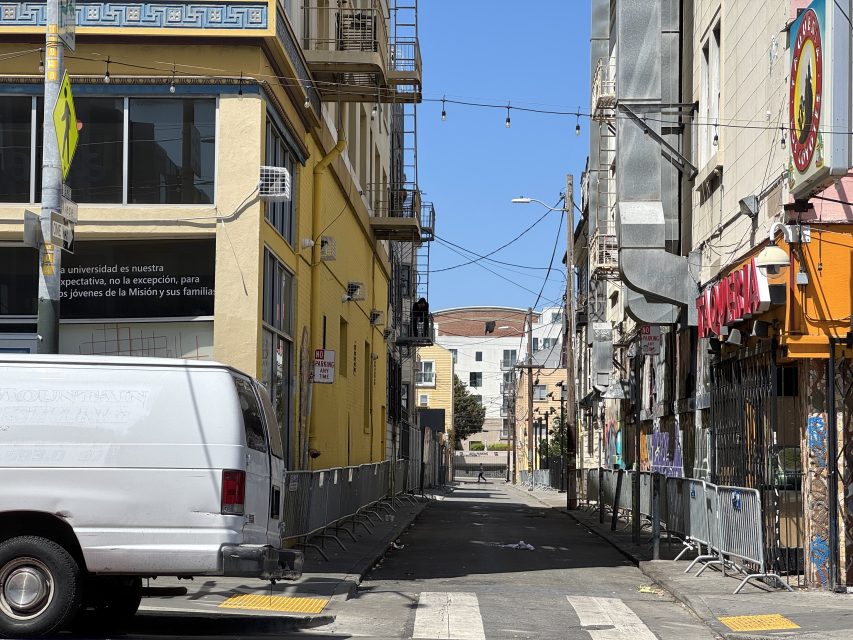 A narrow urban alleyway with buildings on both sides, a white van parked on the left, and string lights hanging above. Fencing lines the alley and a sunny sky is visible.