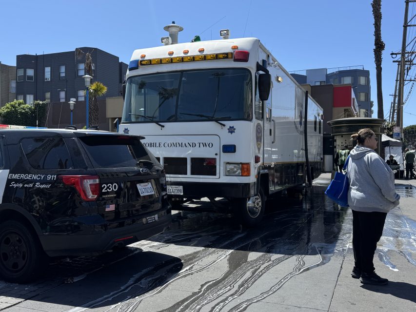 A police SUV and a large mobile command vehicle are parked on a city street while pedestrians walk nearby in daylight.