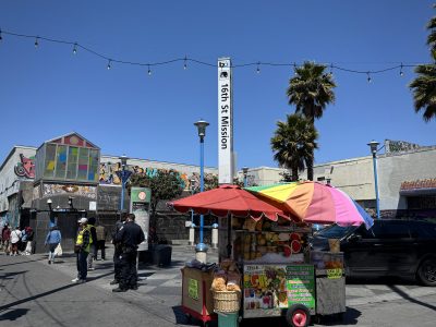 A street vendor cart with snacks and drinks stands near the 16th St Mission BART station sign in a busy urban plaza with people, palm trees, and murals in the background.