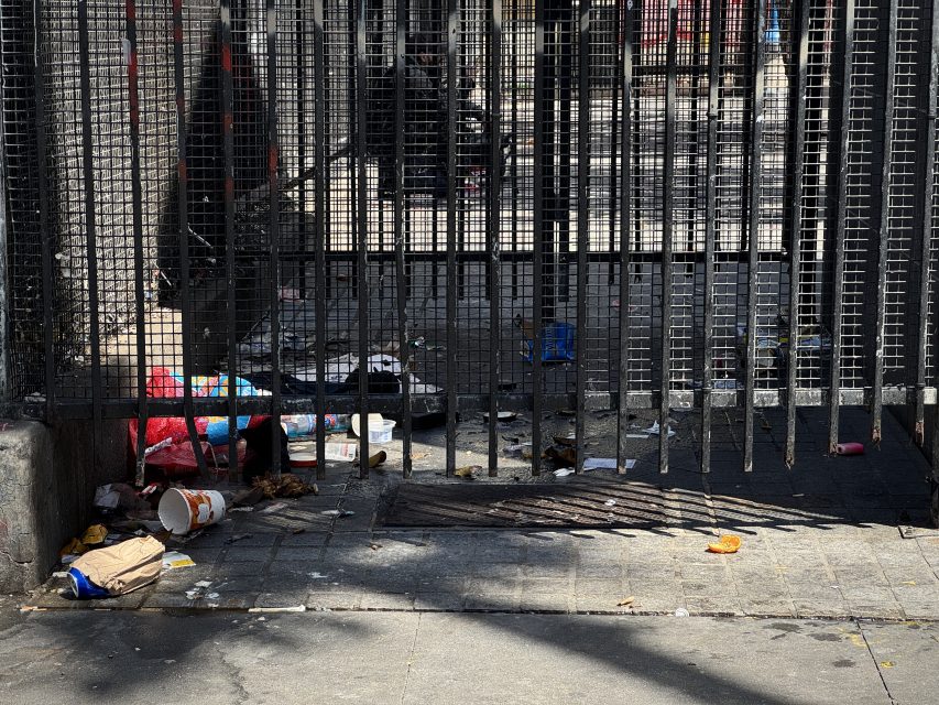 A gated area with metal bars, scattered trash, and debris on the ground, including food containers, paper, and plastic items. Sunlight casts shadows on the pavement.