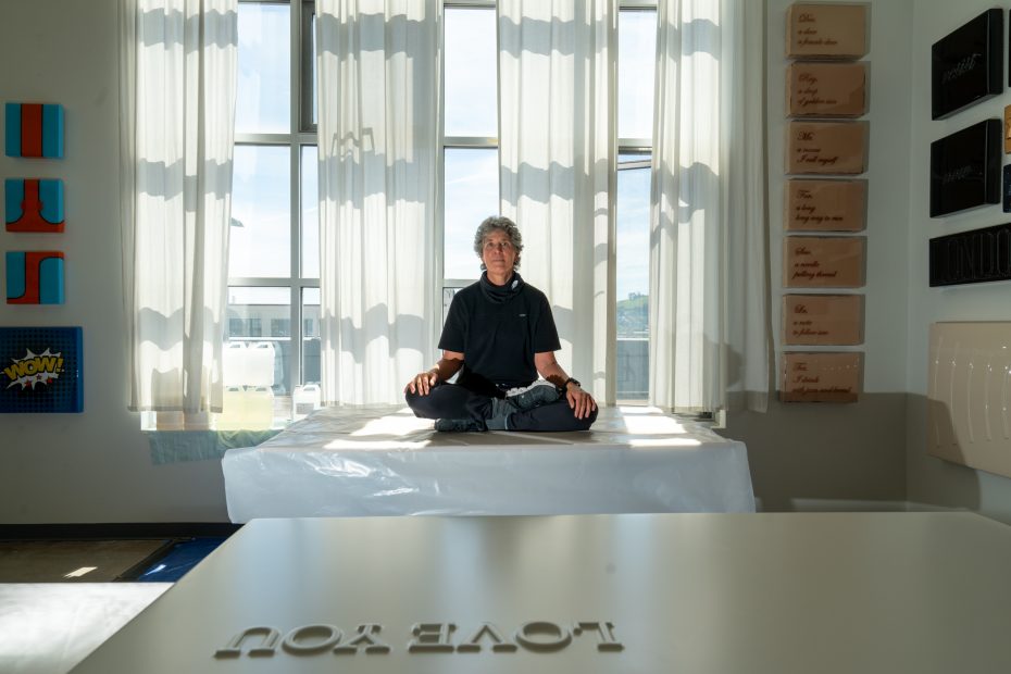 Person sitting cross-legged on a table in a sunlit room with large windows, surrounded by wall art and a reflection of "LOVE YOU" on a surface in the foreground.