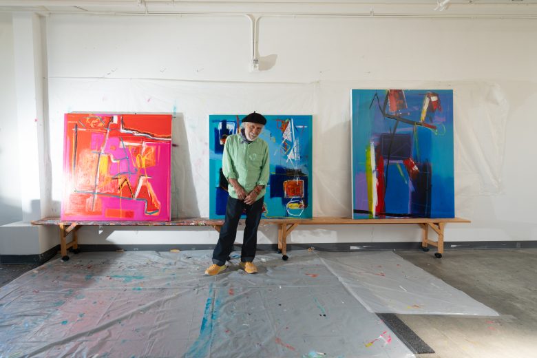 An artist stands in front of three abstract paintings, featuring vibrant colors such as red, pink, and blue, on a bench in a well-lit studio. Plastic sheets cover the floor.