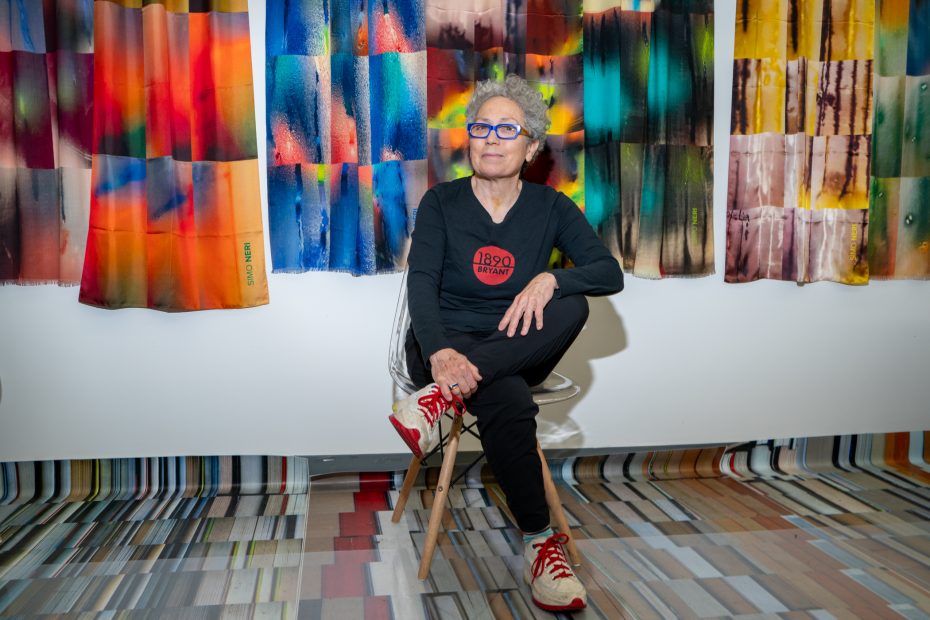 Person with short gray hair sitting in front of colorful abstract fabric panels, wearing glasses, a black shirt with a red logo, black pants, and red sneakers. They have a thoughtful expression.
