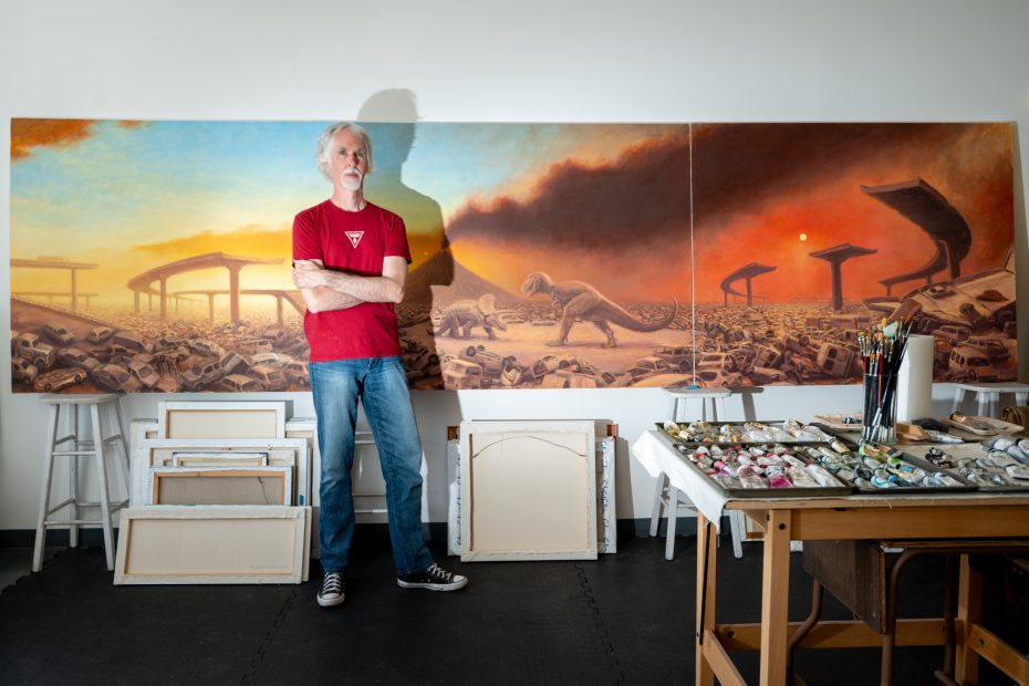 Artist standing with arms crossed in front of large canvas paintings featuring dinosaurs in a post-apocalyptic landscape; art supplies and frames are nearby.