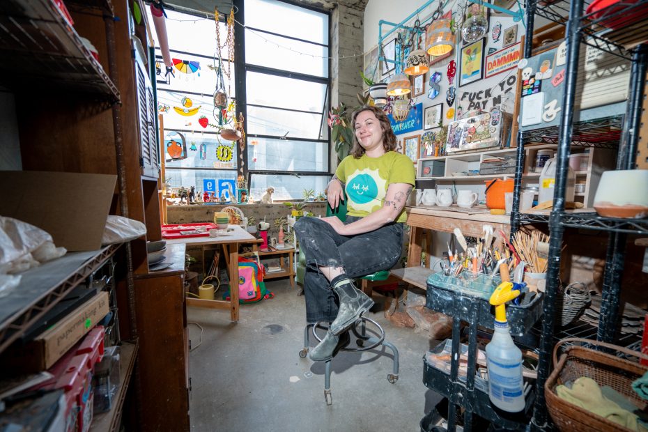 Person sitting on a chair in a cluttered, colorful workshop surrounded by art supplies and decorations, with large windows in the background.
