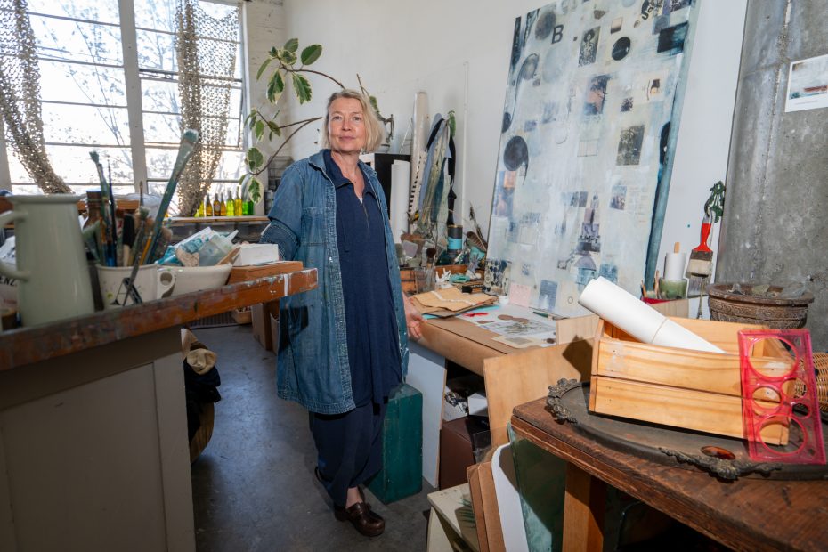 A person stands in an art studio surrounded by various art supplies, a large canvas, and tools.