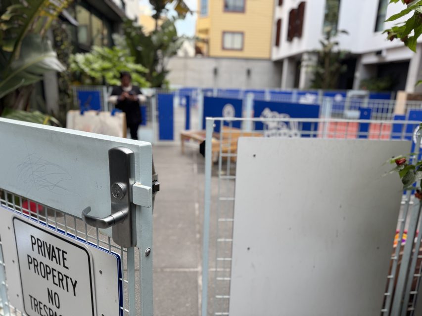 A metal gate with a "Private Property No Trespassing" sign is open, revealing a fenced area with blue panels and a person standing in the background.