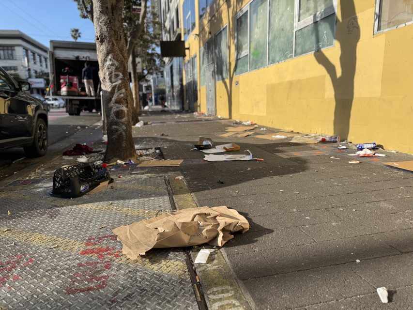Trash and litter are scattered along a city sidewalk next to a yellow building, with a truck and parked cars visible in the background.