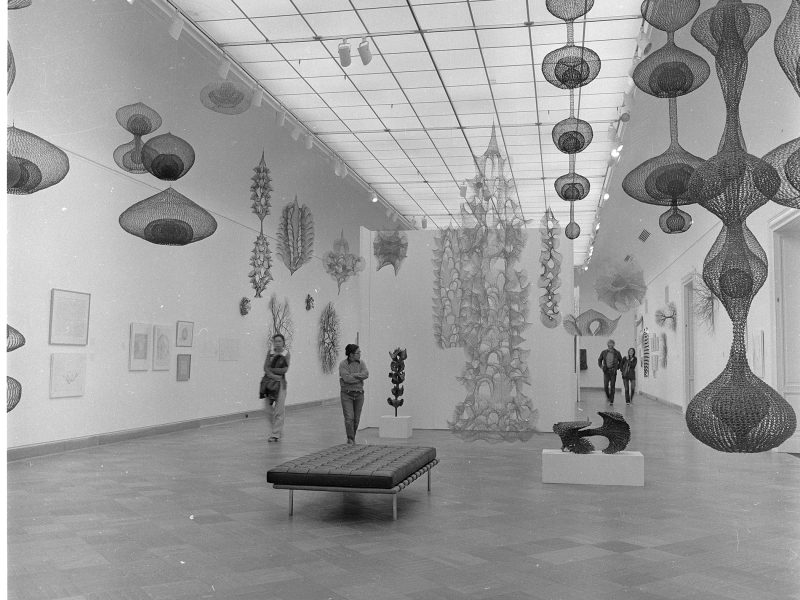 Black and white photo of a gallery with visitors viewing hanging net sculptures and wall art. There's a bench in the center and artworks displayed on pedestals.