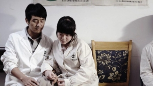 Two people in white lab coats sit closely together on a bench, holding hands, next to an empty chair with a patterned cushion.